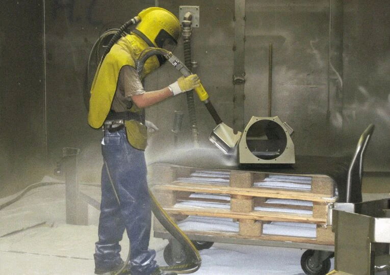 Worker performing sandblasting process on metal casting to remove oxide scale and improve surface roughness in a foundry workshop.